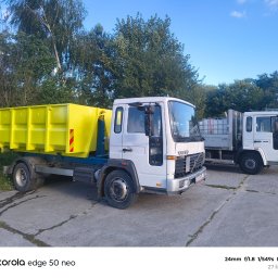 Kiełkujące Kłopoty Rafał Stolarz - Dwie białe ciężarówki Volvo, jedna z jaskrawożółtym, pustym pojemnikiem transportowym, stoją na betonowym placu. W tle zielone drzewa i błękitne niebo.