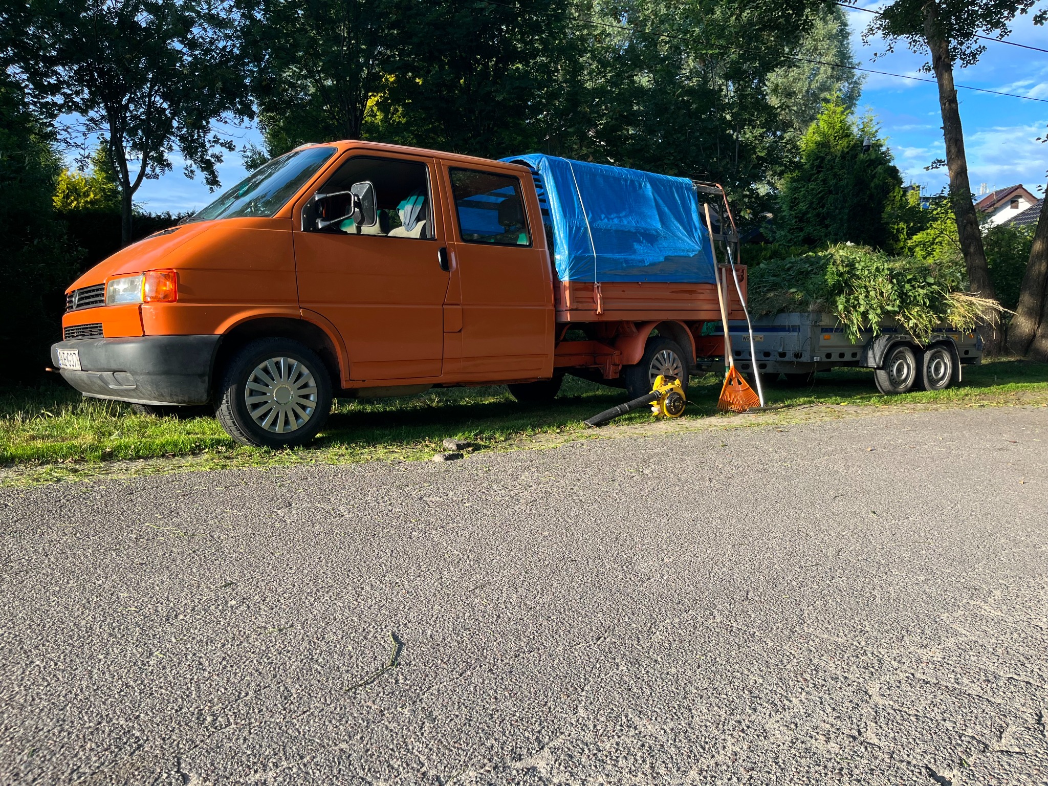 Pomarańczowy samochód dostawczy z przyczepą pełną zieleni, narzędzia ogrodnicze obok, na tle drzew. Wywóz bioodpadów po pielęgnacji roślinności.