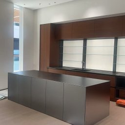 QLdecor - Nowoczesna kuchnia z wyspą ze stali nierdzewnej. Minimalistyczny design, drewniane szafki wiszące, duże okno z widokiem na zewnątrz. Jasna podłoga.