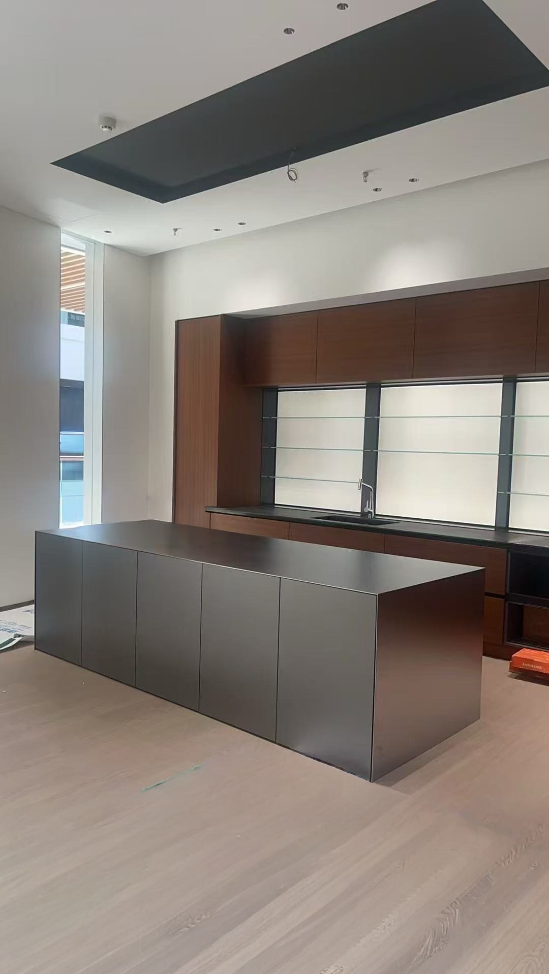 Nowoczesna kuchnia z wyspą ze stali nierdzewnej. Minimalistyczny design, drewniane szafki wiszące, duże okno z widokiem na zewnątrz. Jasna podłoga.