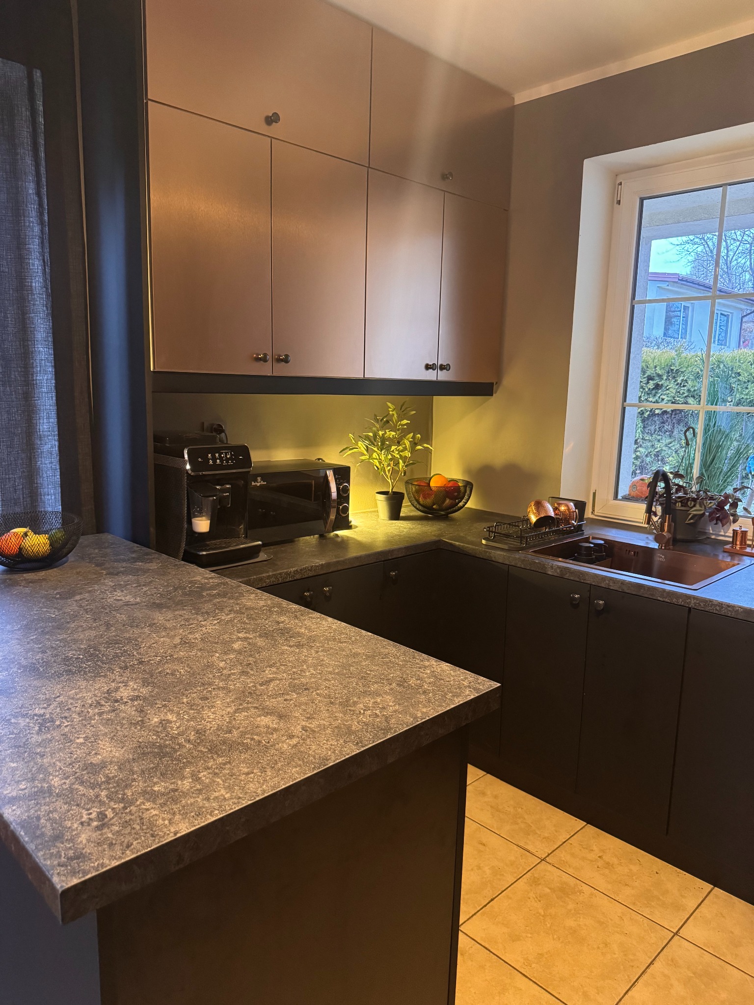 Nowoczesna kuchnia z metalizowanymi frontami w odcieniach miedzi, ciemny blat i zabudowa, widok na okno z roślinami. Ekspres do kawy i mikrofalówka na blacie.