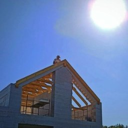 DeWuConstruction - budowa domów pasywnych i realizacji pod klucz - Budowa domu w stanie surowym otwartym w słoneczny dzień. Widoczna konstrukcja dachu z drewnianymi belkami i ściany z betonu komórkowego. Na szczycie dachu stoi osoba.