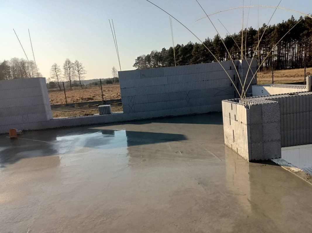 Wylana, gładka posadzka w surowym stanie, z widocznymi fragmentami ścian z szarych bloczków. W tle drzewa i błękitne niebo. Widok na plac budowy w słoneczny dzień.