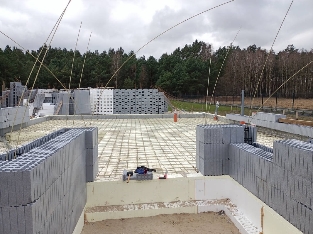 Budowa domu: fundamenty z bloczków betonowych, zbrojenie siatką stalową, w tle las i ułożone bloczki. Widoczne narzędzia budowlane.