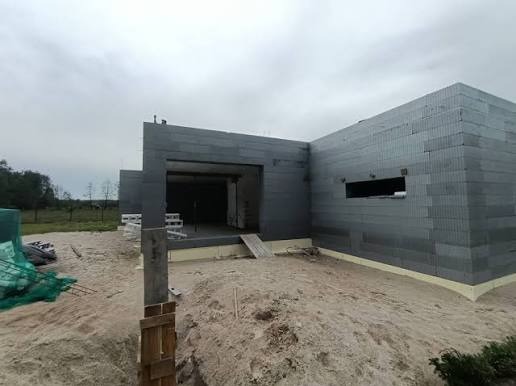 Surowy stan domu z szarej cegły, widok z zewnątrz na plac budowy z piaskiem i materiałami budowlanymi. Widoczne otwory okienne i drzwiowe.