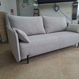 Tapicerstwo Meblowe - Jasnoszara sofa dwuosobowa z poduszkami, tapicerowana tkaniną o wyraźnej fakturze, na tle warsztatu z widocznym znakiem zakazu palenia.