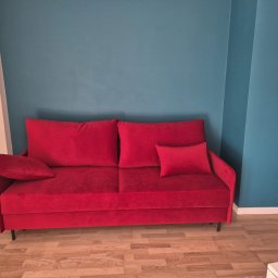 Tapicerstwo Meblowe - Czerwona, welurowa sofa z poduszkami na tle turkusowej ściany. Minimalistyczny design, czarne nóżki. Widok z przodu, parkiet na podłodze.