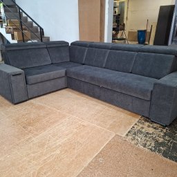 Tapicerstwo Meblowe - Szara, narożna sofa tapicerowana z podnoszonym siedziskiem, widoczna w warsztacie na tle schodów i materiałów. Nowoczesny design, proste linie, ciemna tkanina.