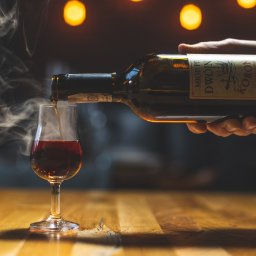 Photostories - Nalewanie miodu pitnego 'Dwójniak Koronny' z butelki do kieliszka na drewnianym blacie, z subtelnym dymem i ciepłym oświetleniem w tle.