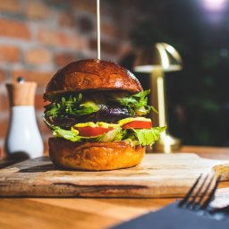 Photostories - Apetyczny burger z warzywami na drewnianej desce, w tle ceglana ściana i przyprawniki. Oświetlenie podkreśla świeżość składników.