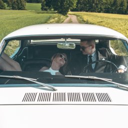 Photostories - Para w okularach przeciwsłonecznych w białym, klasycznym samochodzie na tle zielonego pola i drogi. Kobieta z nogami wystawionymi przez okno.