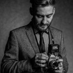 Photostories - Czarno-białe zdjęcie fotografa w tweedowej marynarce, z brodą, spoglądającego na aparat Praktica z lampą błyskową Toshiba 300, na szarym tle.