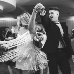 Photostories - Czarno-białe zdjęcie pary w tańcu na parkiecie podczas uroczystości. Kobieta w sukience z frędzlami, mężczyzna w garniturze. Dynamiczne ujęcie, rozmazane tło.