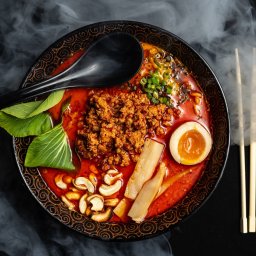 Photostories - Aromatyczny ramen w czarnej misce z wzorem, z jajkiem, grzybami, mięsem i zielenią, otoczony subtelną mgłą, z czarną łyżką i pałeczkami. Zdjęcie z góry.