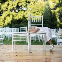 Photostories - Dziewczynka w białej sukience opiera się o białe krzesła na drewnianym podeście, w tle rząd białych krzeseł i zielone drzewa. Uwagę przykuwają srebrne buciki.