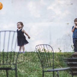 Photostories - Dzieci na tle białej ściany: dziewczynka w czarnej sukience z pomarańczowym balonem, chłopiec w niebieskiej koszuli. W tle trawa i krzesła.
