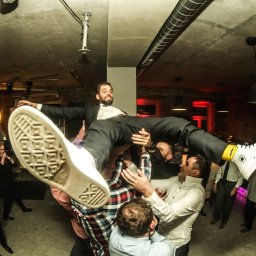 Photostories - Uśmiechnięty mężczyzna w garniturze, podrzucany do góry przez grupę osób podczas imprezy. Widoczne buty Converse i surowy wystrój wnętrza.