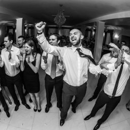 Photostories - Czarno-białe zdjęcie z imprezy. Grupa osób w formalnych strojach świętuje, mężczyzna w centrum z podniesioną ręką i otwartymi ustami. Widoczny parkiet i dekoracje.