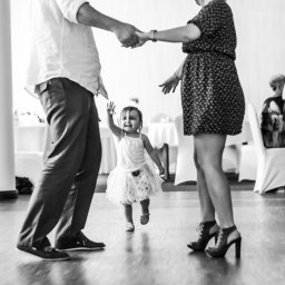 Photostories - Radosna dziewczynka w tiulowej sukience biegnie w stronę aparatu, trzymając za ręce rodziców na parkiecie podczas przyjęcia. Czarno-biała fotografia z gośćmi w tle.