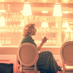 Photostories - Kobieta w sukience w grochy siedzi w barze, pijąc drinka. W tle widoczne oświetlenie i szklanki. Ujęcie z profilu, ciepłe światło, klimatyczne wnętrze.