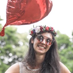 Photostories - Uśmiechnięta kobieta w okularach i wianku, z odbiciem fotografa w szkłach, trzymająca czerwony balon na tle zieleni. Portret w plenerze.