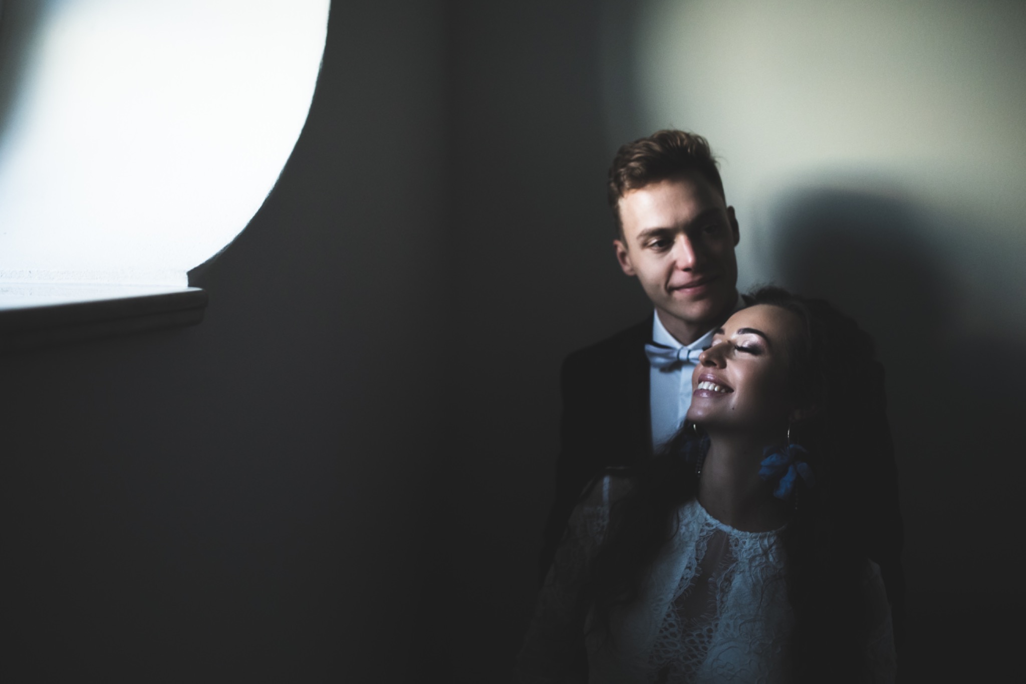 Elegancka para w ciemnym wnętrzu, on w smokingu, ona w koronkowej sukience, subtelne oświetlenie podkreśla emocje. Sesja w Kielcach.