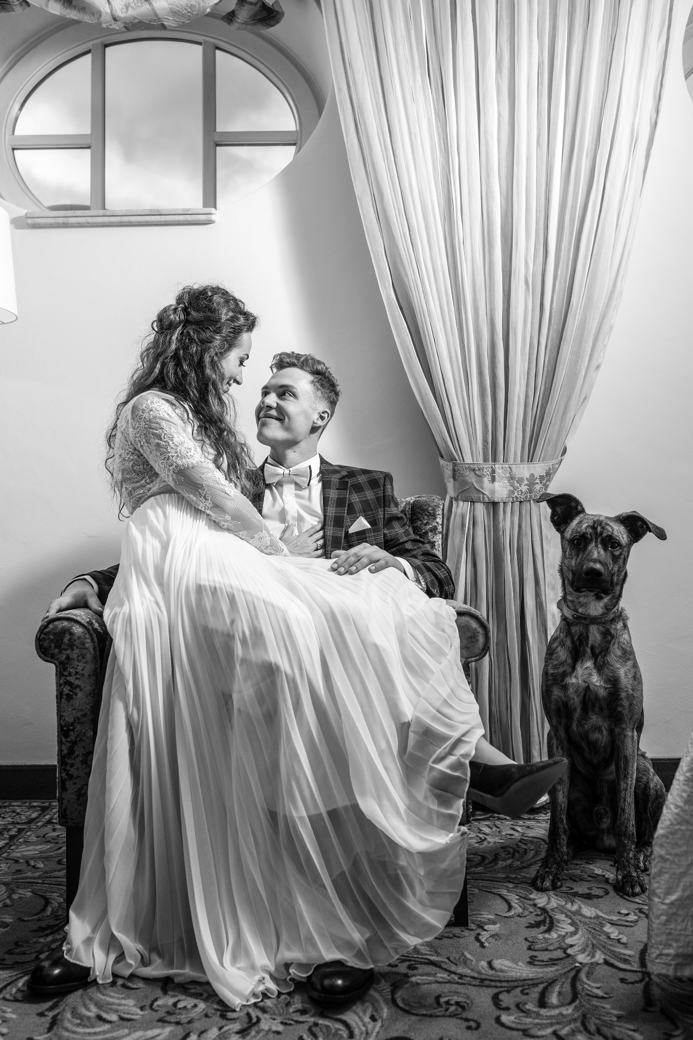 Czarno-białe zdjęcie pary młodej siedzącej w fotelu z psem. Panna młoda w sukni z koronkową górą, pan młody w kraciastej marynarce. Romantyczny nastrój, eleganckie wnętrze.