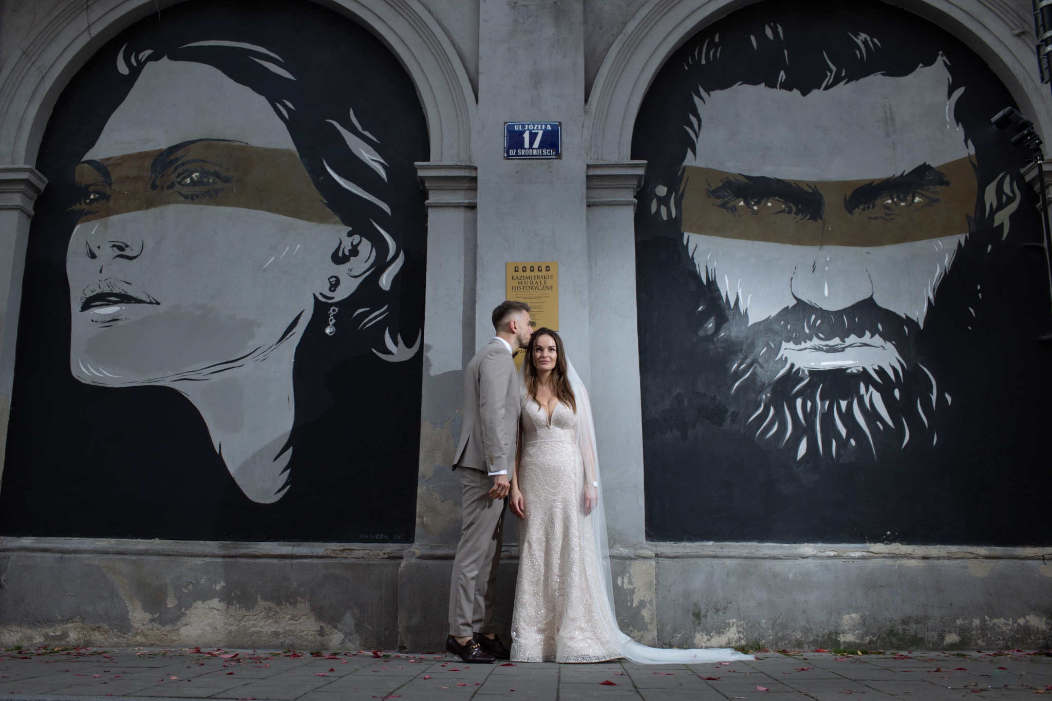 Para młoda w beżowych strojach pozuje na tle ściany z muralami w odcieniach szarości. Pan młody całuje pannę młodą w czoło. Ulica Józefa 17 w tle.
