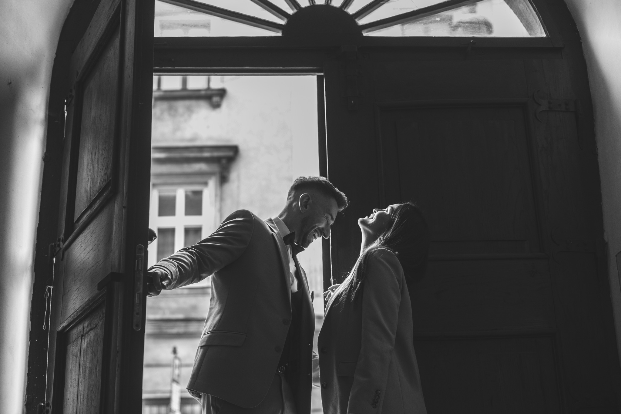 Czarno-białe zdjęcie pary w garniturach, stojącej w otwartych drzwiach z dekoracyjnym nadprożem, skierowanej twarzami ku sobie i uśmiechniętej.