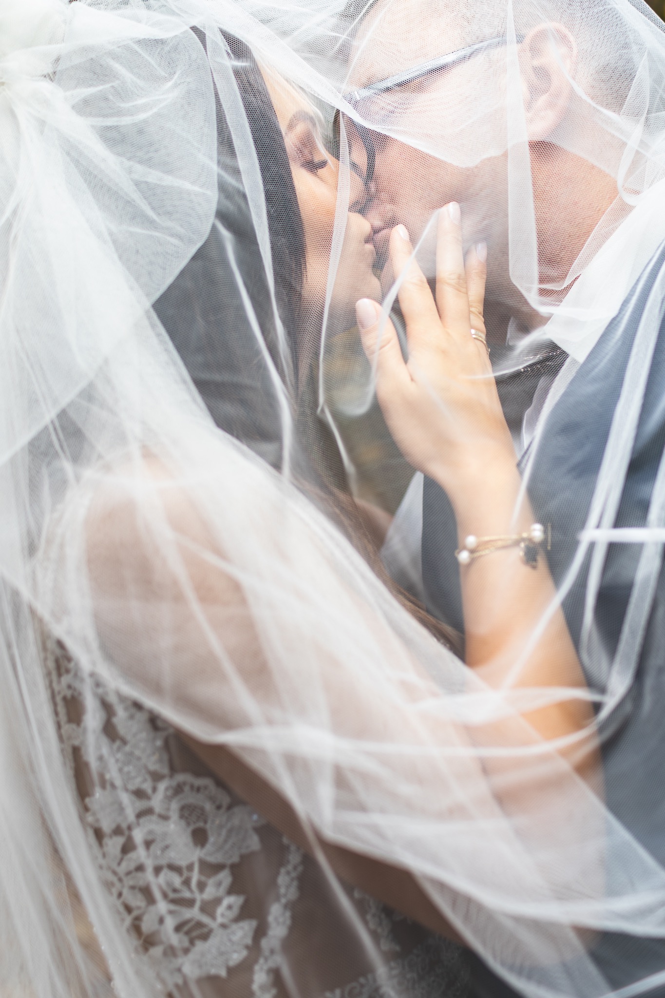 Delikatne ujęcie pocałunku pary młodej pod welonem. Widoczna dłoń panny młodej z pierścionkiem i bransoletką. Subtelne światło podkreśla intymność chwili.