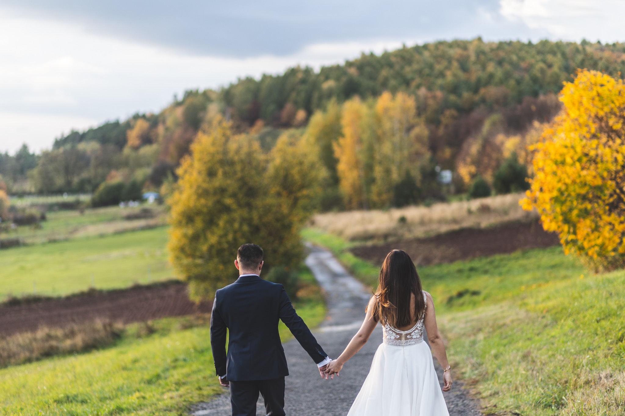 Para młoda, trzymając się za ręce, idzie polną drogą w jesiennej scenerii. Suknia panny młodej z koronkowym tyłem, garnitur pana młodego. Widok z tyłu, ciepłe światło.
