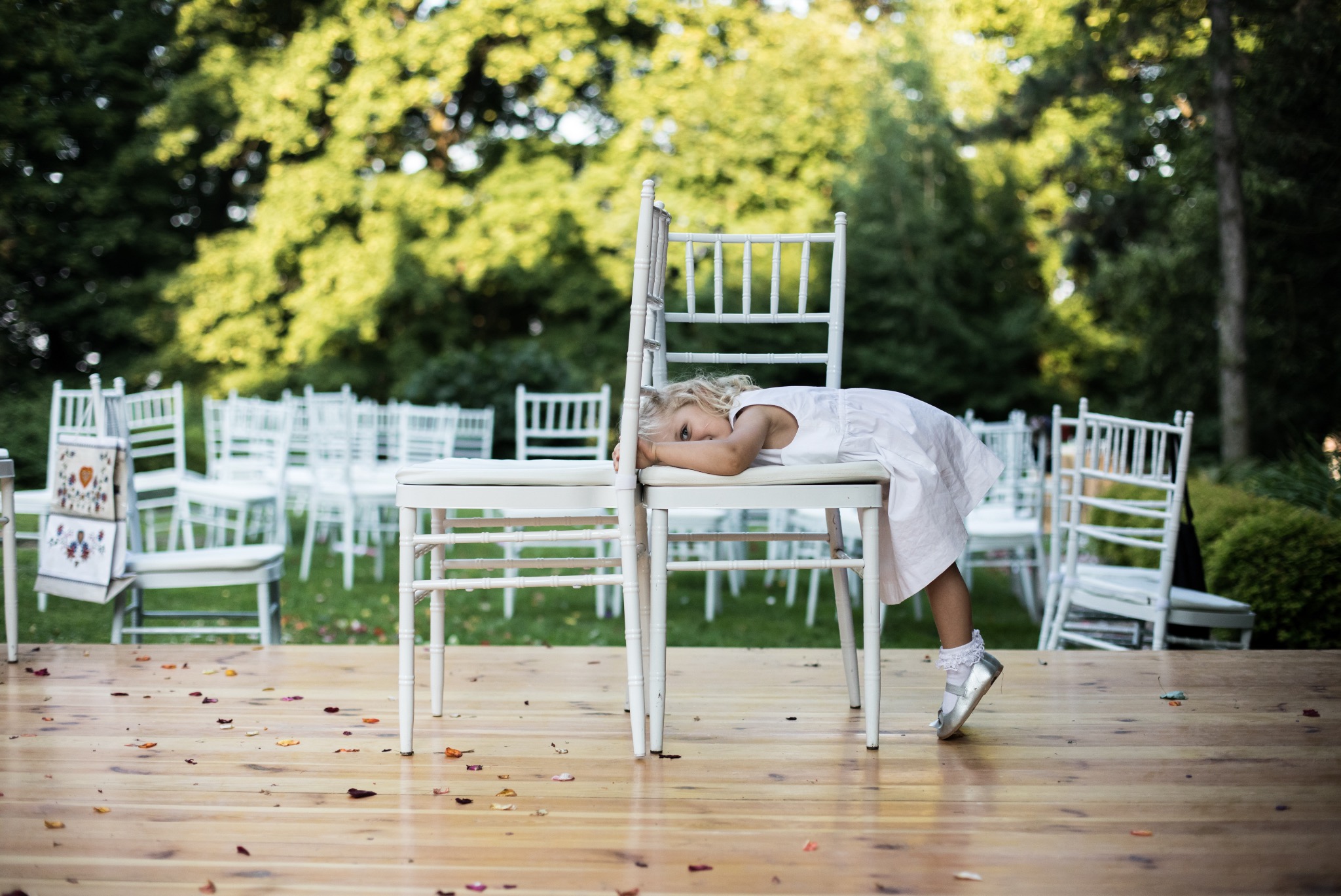 Dziewczynka w białej sukience opiera się o białe krzesła na drewnianym podeście, w tle rząd białych krzeseł i zielone drzewa. Uwagę przykuwają srebrne buciki.