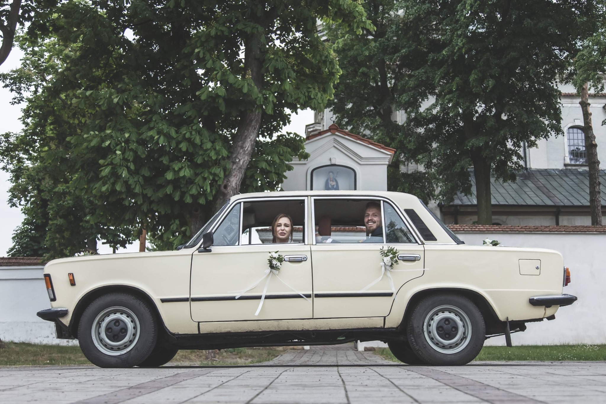 Para młoda w beżowym Fiacie 125p, udekorowanym kwiatami i wstążkami, na tle zieleni i kapliczki. Uśmiechnięci, patrzą w obiektyw.
