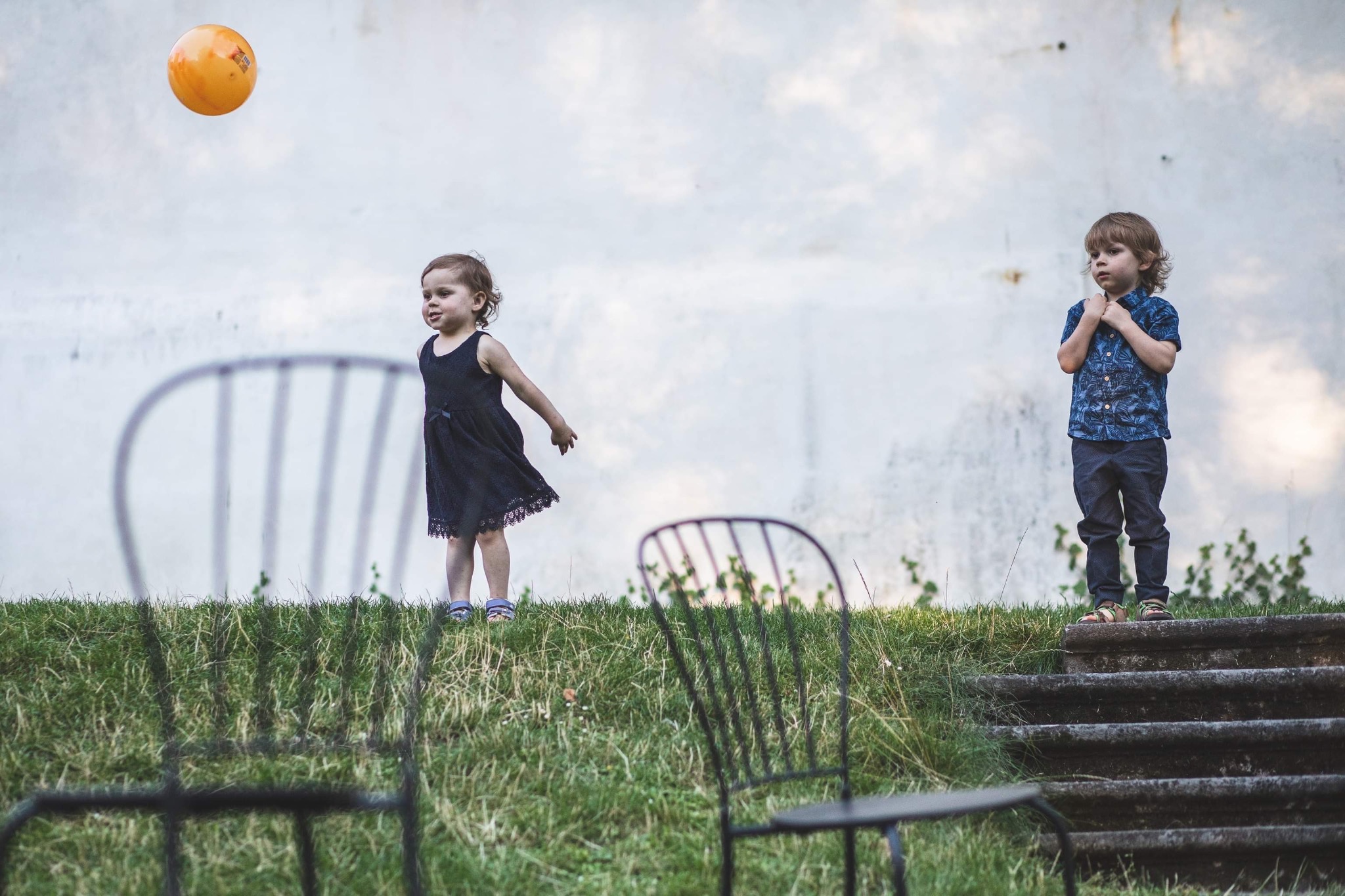 Dzieci na tle białej ściany: dziewczynka w czarnej sukience z pomarańczowym balonem, chłopiec w niebieskiej koszuli. W tle trawa i krzesła.