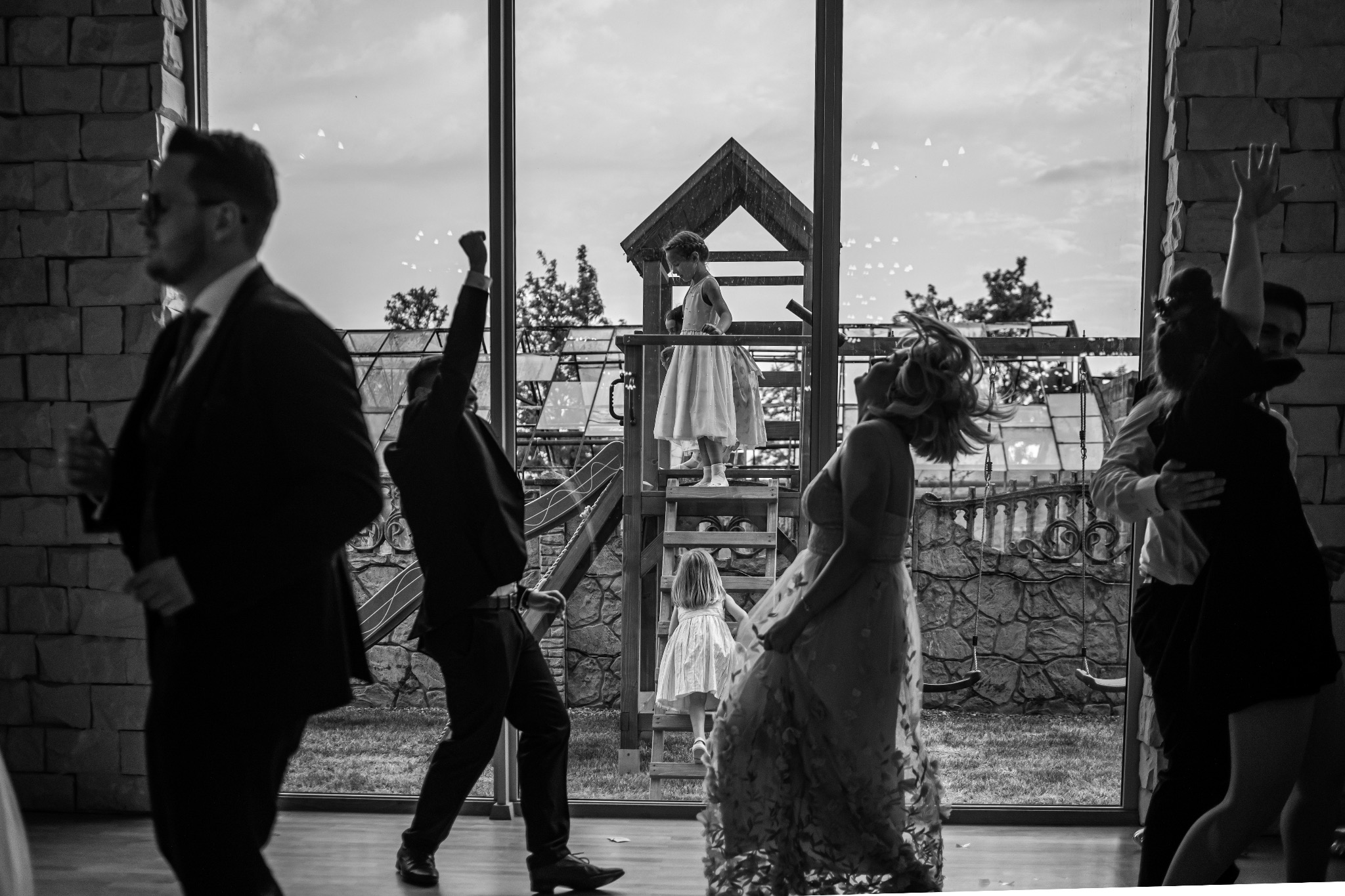Czarno-białe zdjęcie z wesela: goście tańczą w sali z dużym oknem, za którym widać dzieci bawiące się na placu zabaw. Dynamiczne ujęcie, uchwycone emocje.