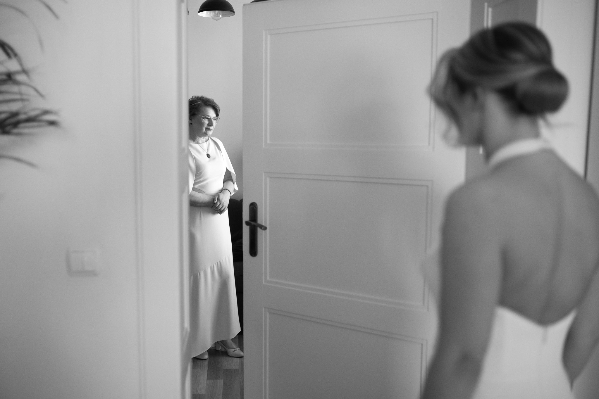 Czarno-białe zdjęcie panny młodej z tyłu, w tle matka w białej sukni, stojąca w drzwiach. Ujęcie zza ramienia, miękkie światło, przygotowania do ślubu.