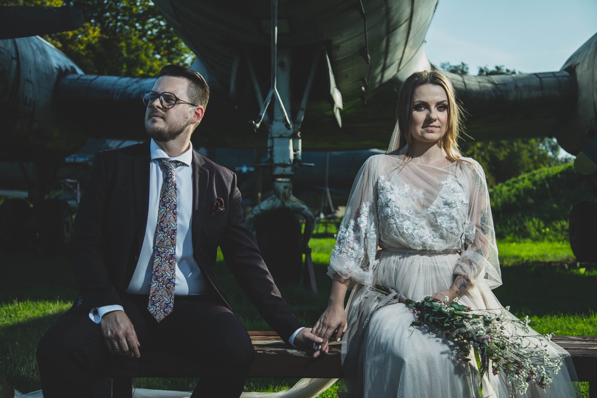 Para młoda siedzi na ławce przed starym samolotem. Pan młody w bordowym garniturze i wzorzystym krawacie, panna młoda w zwiewnej sukni trzyma bukiet. Nietypowa sesja ślubna.