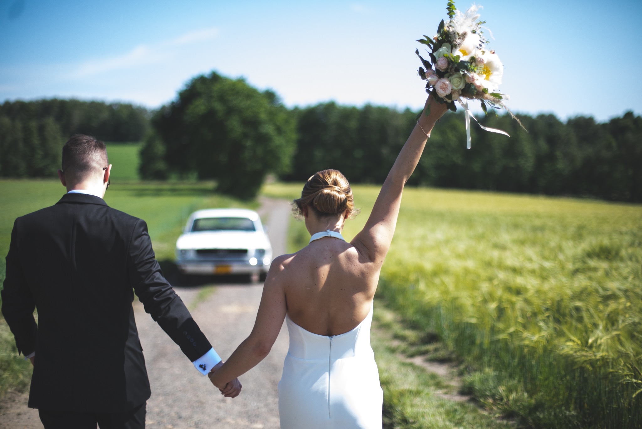 Para młoda, trzymając się za ręce, idzie polną drogą w stronę białego samochodu. Panna młoda unosi bukiet kwiatów. Ujęcie z tyłu, w słoneczny dzień.
