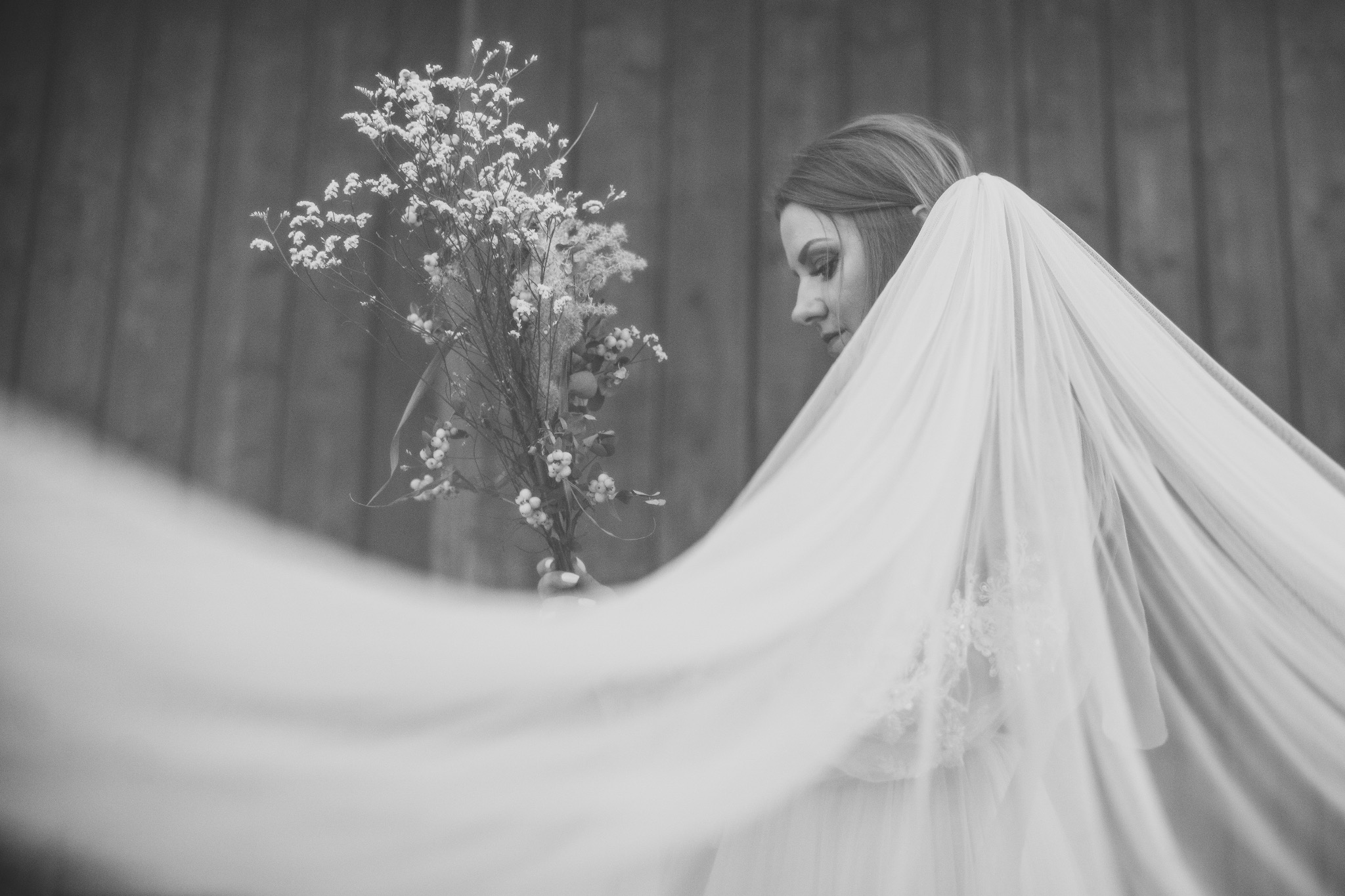 Monochromatyczne ujęcie panny młodej z bukietem polnych kwiatów, zwiewnym welonem i delikatnym makijażem na tle drewnianej ściany. Profil, subtelność, naturalność.