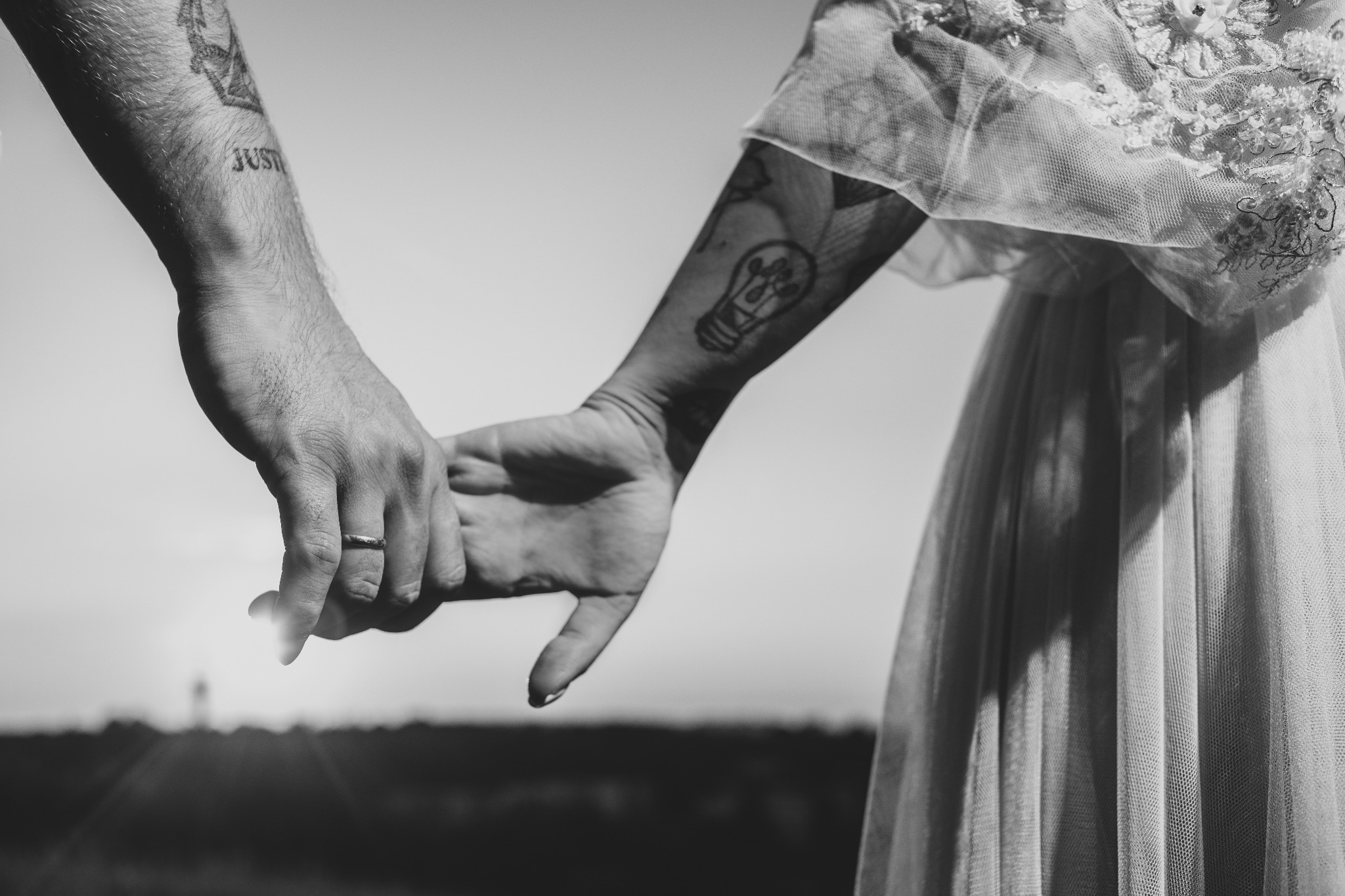Czarno-białe zbliżenie dłoni pary z tatuażami i obrączką, trzymających się na tle zachodzącego słońca i sukni ślubnej. Romantyczny kadr z plenerowej sesji.