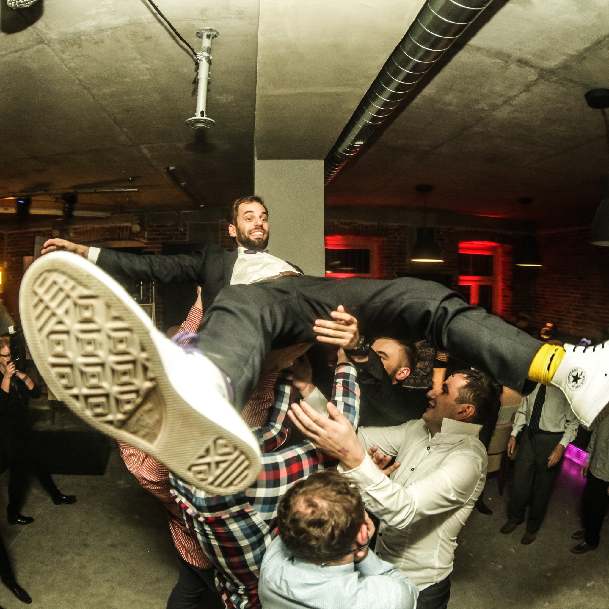 Uśmiechnięty mężczyzna w garniturze, podrzucany do góry przez grupę osób podczas imprezy. Widoczne buty Converse i surowy wystrój wnętrza.