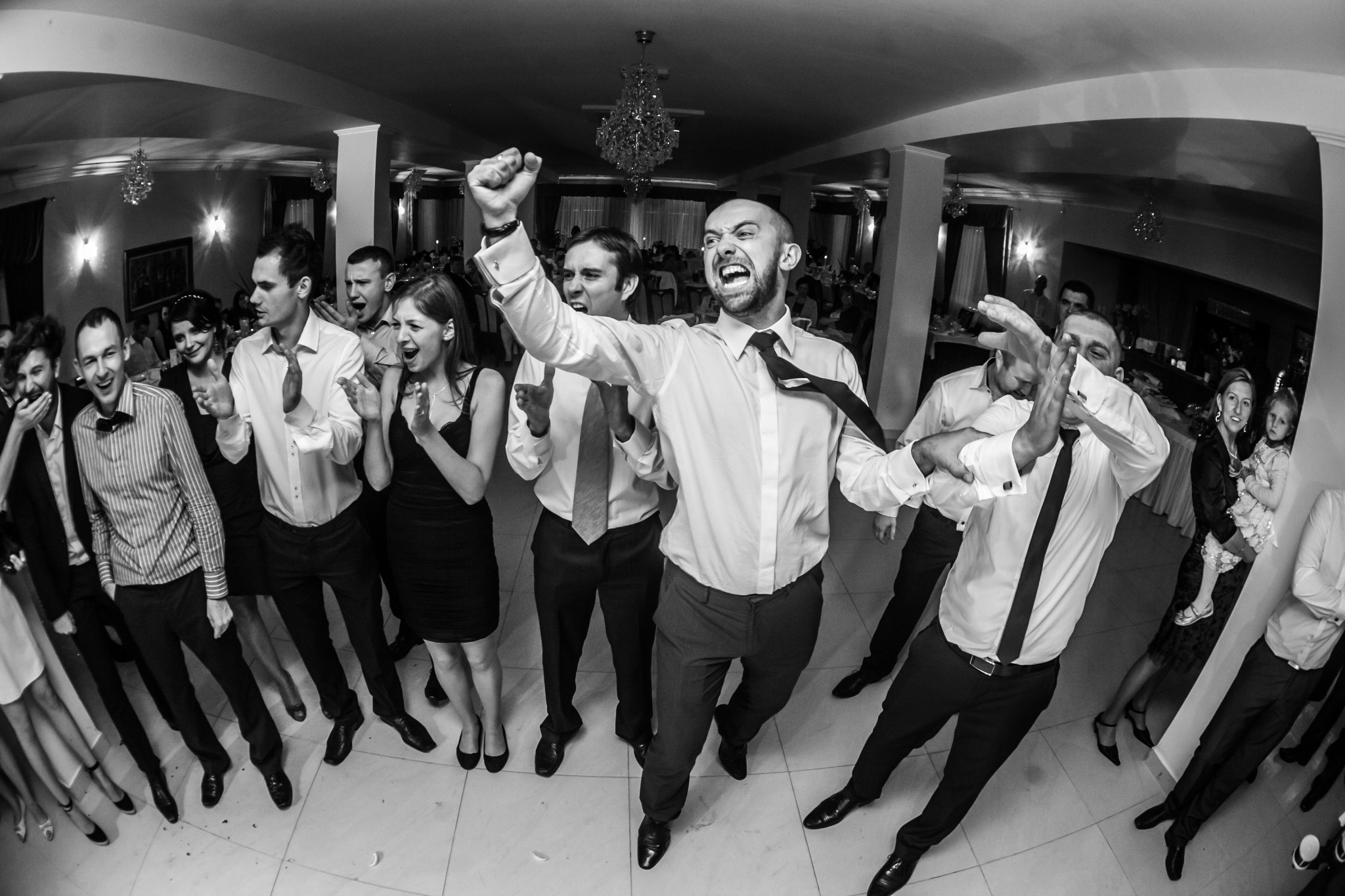 Czarno-białe zdjęcie z imprezy. Grupa osób w formalnych strojach świętuje, mężczyzna w centrum z podniesioną ręką i otwartymi ustami. Widoczny parkiet i dekoracje.