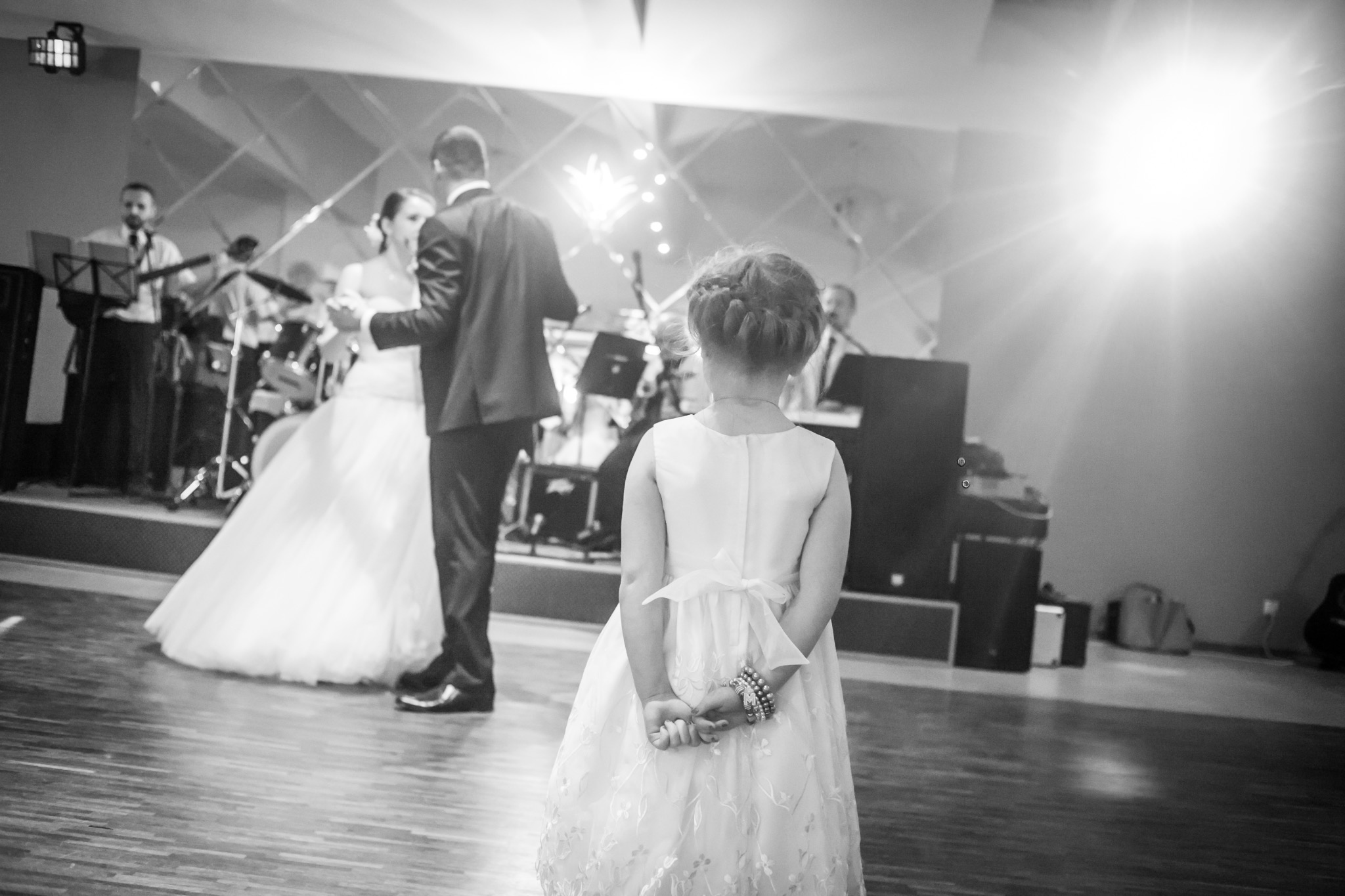 Czarno-białe zdjęcie dziewczynki w sukience, obserwującej pierwszy taniec pary młodej z zespołem muzycznym w tle, uchwycone zza jej pleców.