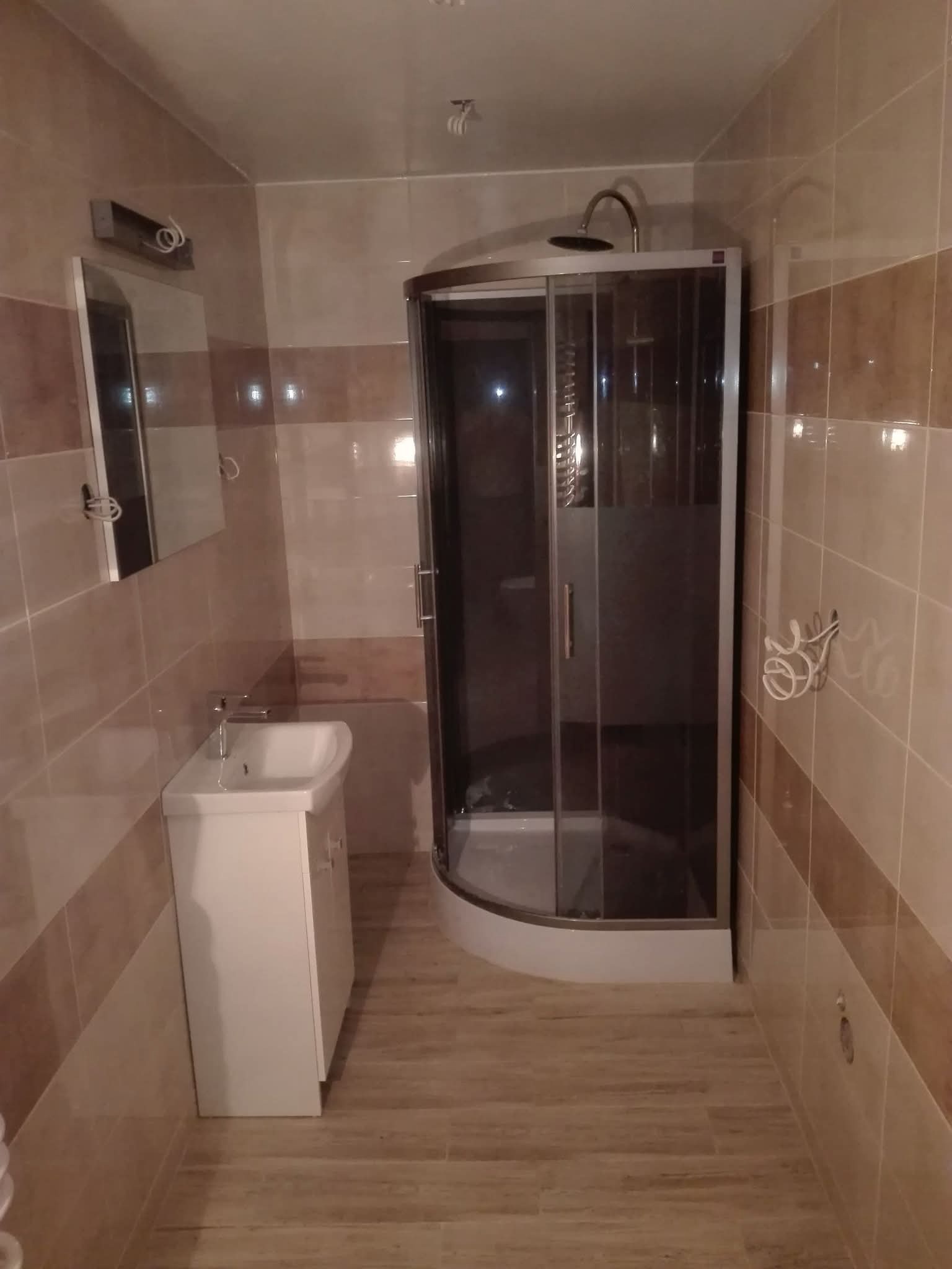 Wykończona łazienka z kabiną prysznicową, umywalką z szafką i lustrem. Płytki w odcieniach beżu i brązu. Widoczne przewody elektryczne na ścianie.