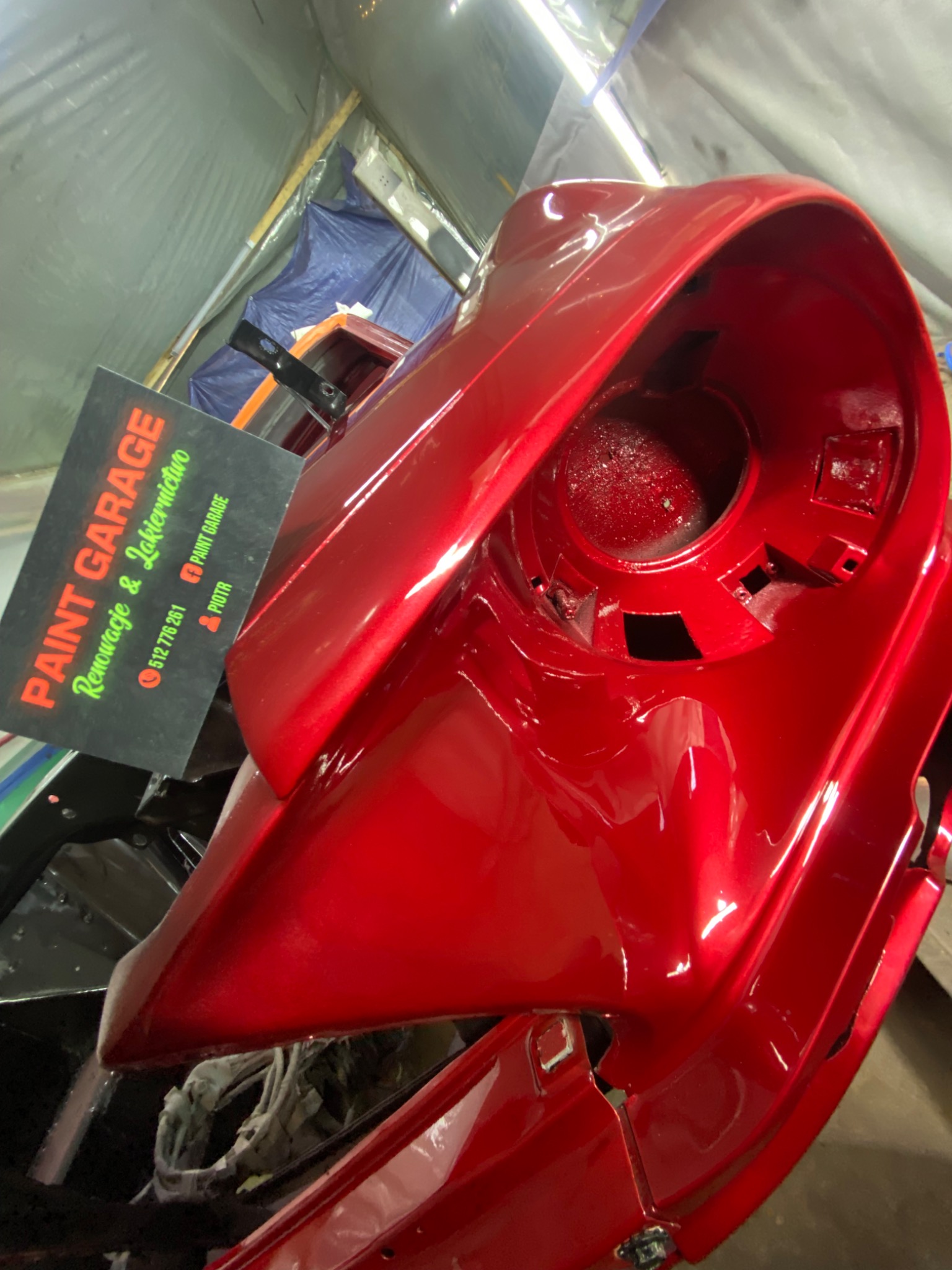 Czerwony element karoserii auta po lakierowaniu, z połyskiem, w warsztacie. Obok wizytówka 'Paint Garage' z danymi kontaktowymi. Nietypowy kadr z bliska.