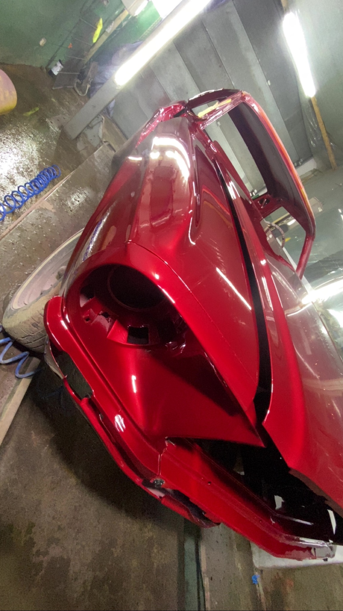 Czerwony element karoserii samochodu po lakierowaniu, z widocznym połyskiem świeżej farby, w warsztacie z oświetleniem jarzeniowym i widocznym przewodem pneumatycznym.