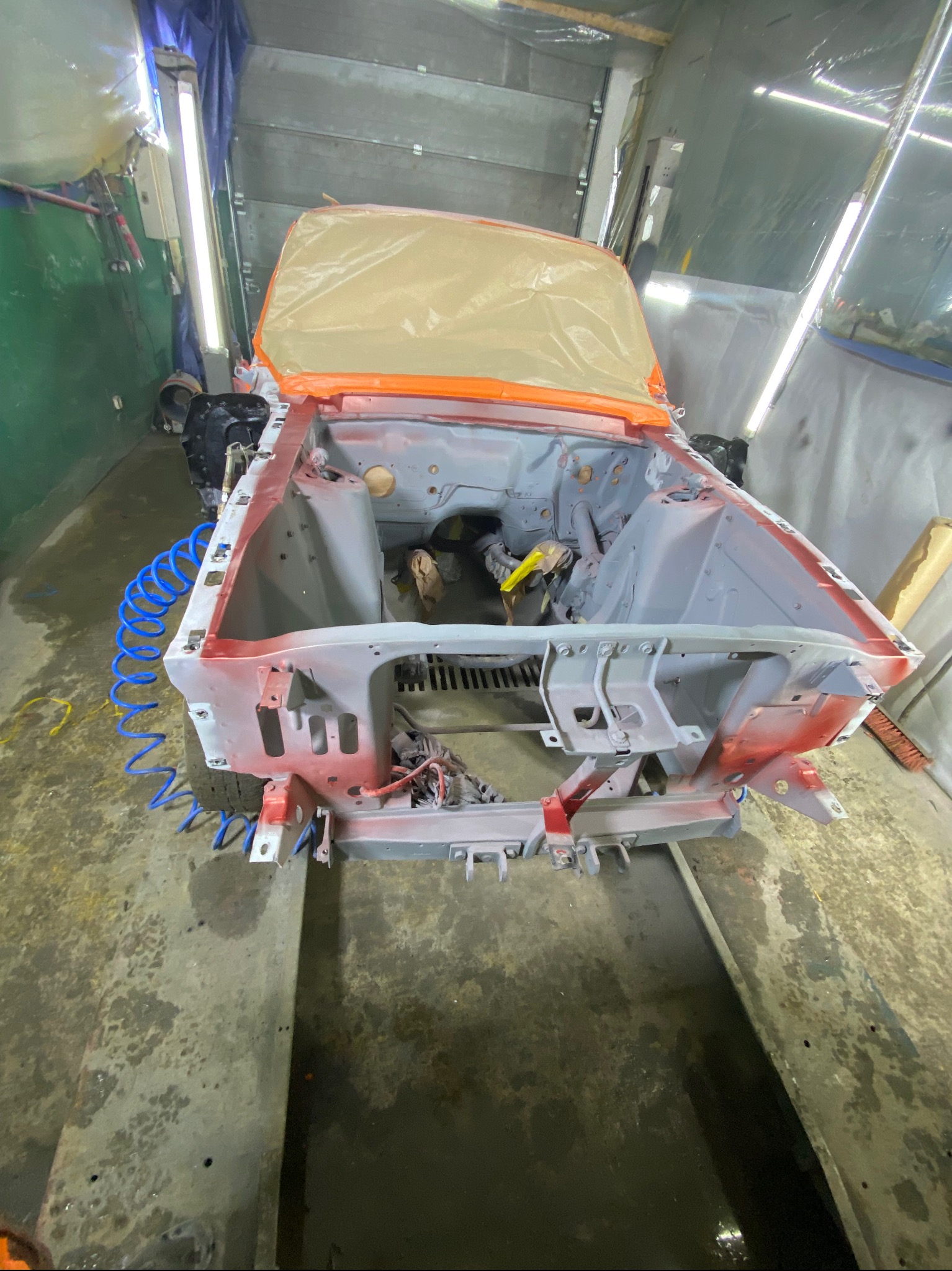 Karoseria samochodu w trakcie renowacji, zabezpieczona folią, z widocznymi śladami szpachli i podkładu, w warsztacie z niebieskim przewodem pneumatycznym.