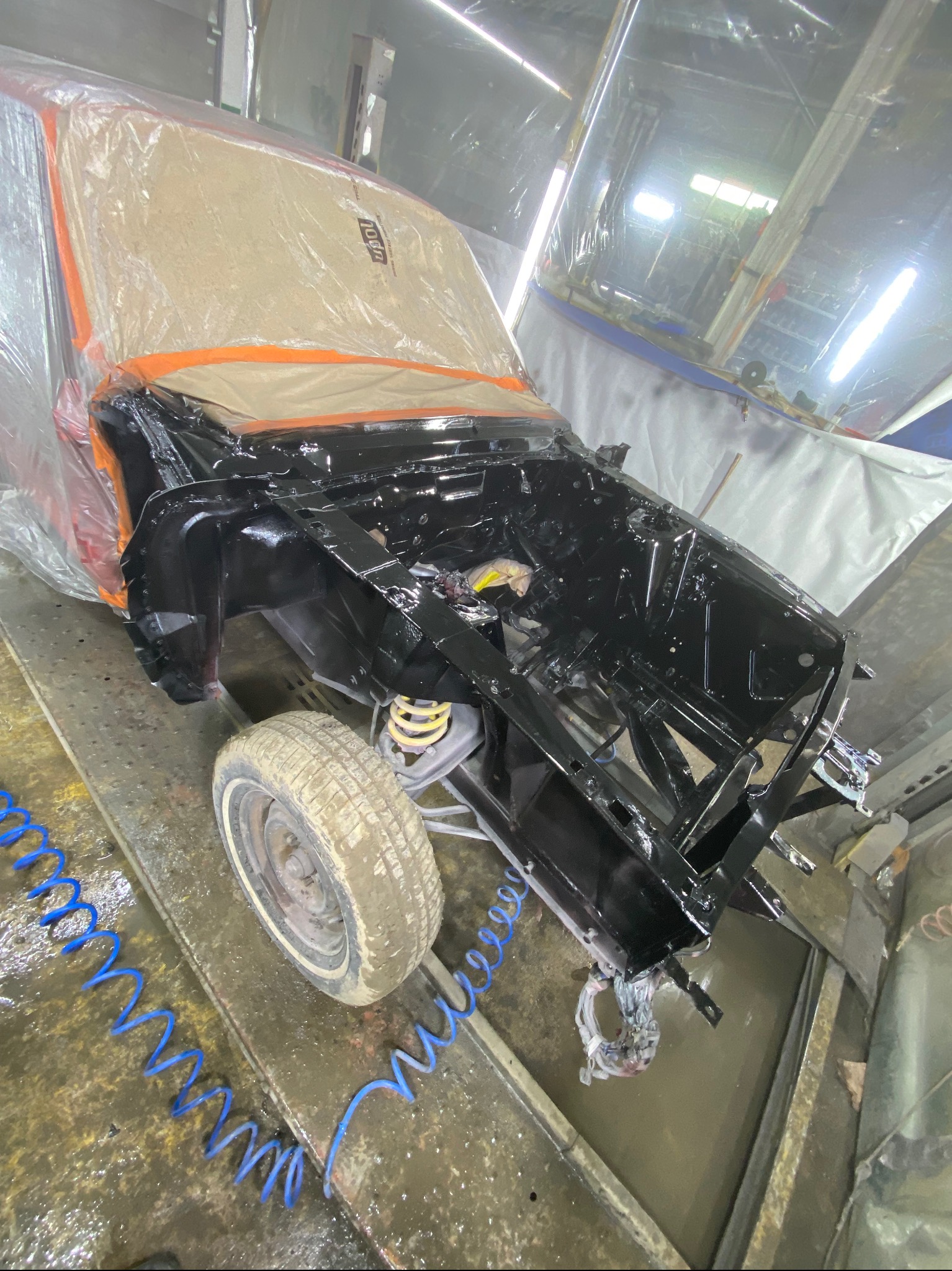 Częściowo zdemontowany, czarny samochód w trakcie lakierowania, zabezpieczony folią i taśmą. Widoczne elementy zawieszenia i koło. Podłoże mokre, z przewodami.