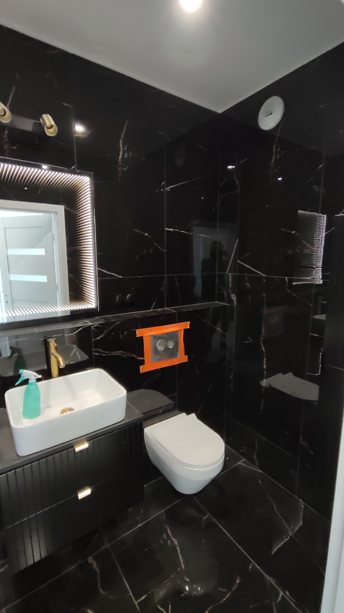 Elegancka, czarno-biała łazienka z marmurowymi płytkami, podświetlanym lustrem, białą umywalką na szafce i wiszącą toaletą. Widoczny fragment instalacji podtynkowych.