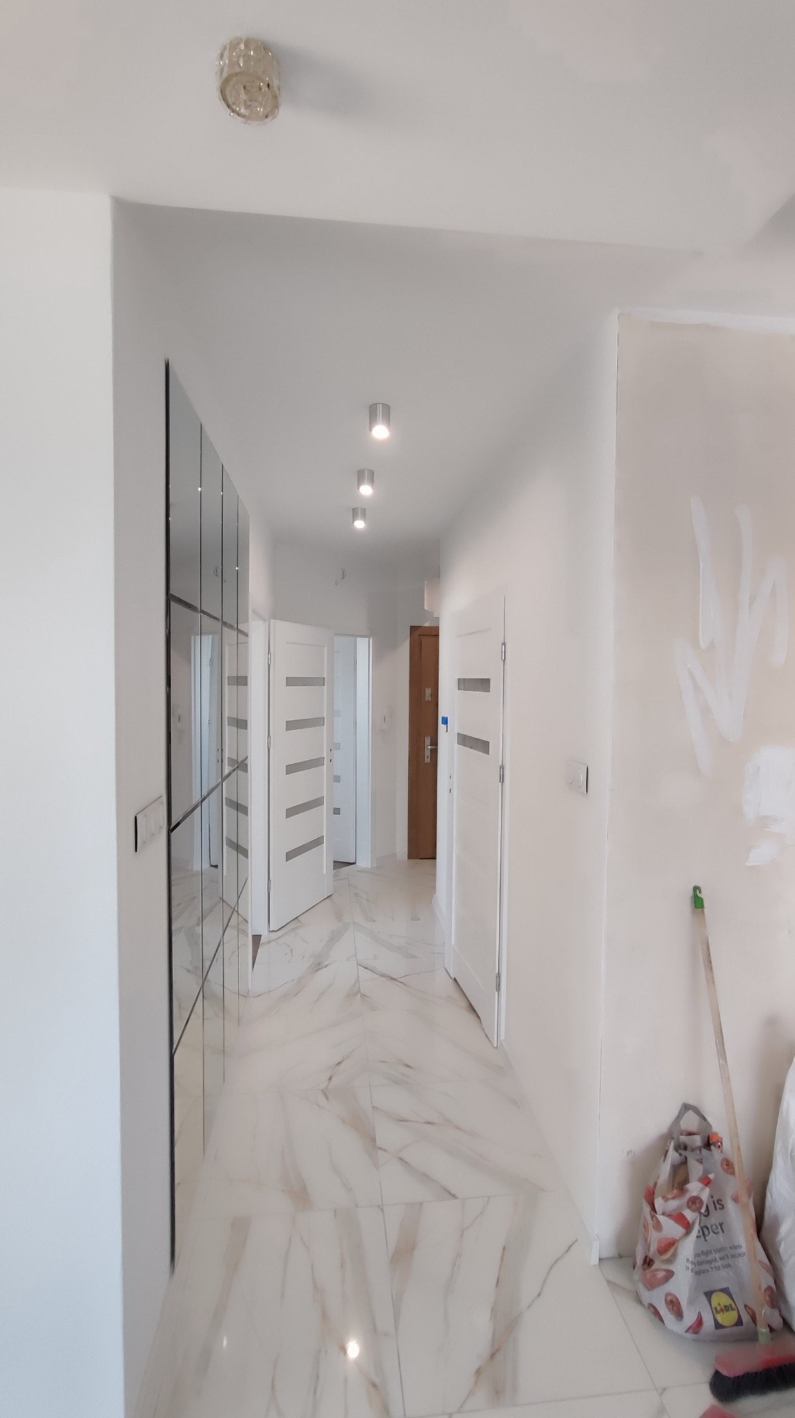 Biały korytarz z marmurową podłogą i lustrzaną szafą. Widoczne drzwi wewnętrzne z poziomymi przeszkleniami i ślady po gładzi na ścianie.